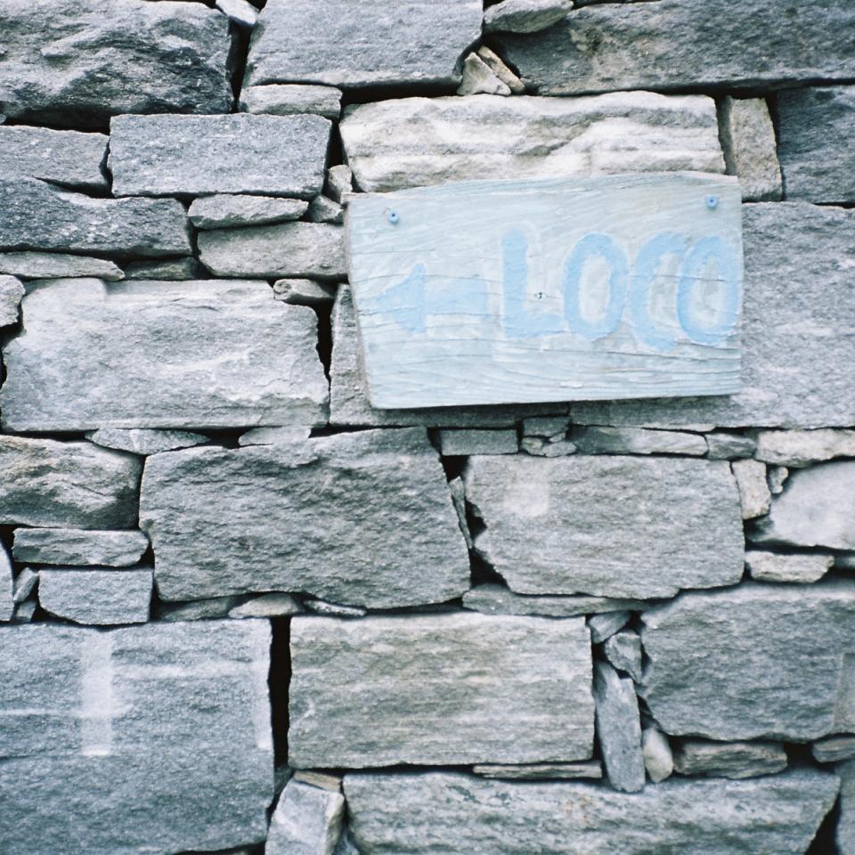 Cover Photography portraying a natural stone house wall with a wooden sign saying "←Loco"