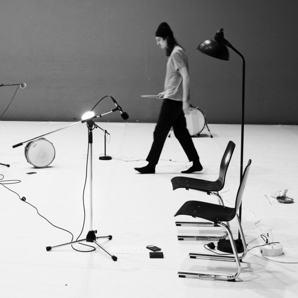 Photo of Paul Amereller walking through a space with chairs, mic stand and drums on the floor, holding a drum stick