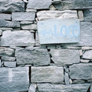 Cover Photography portraying a natural stone house wall with a wooden sign saying "←Loco"